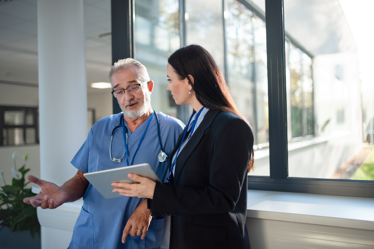 A health care manager talks with a physician wearing scrubs in a hospital corridor.