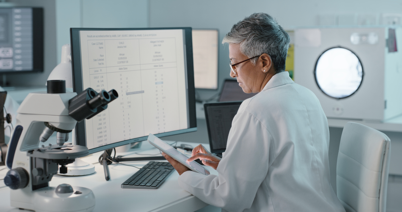 A forensic science technician works at a desk with a tablet, PC and microscope.