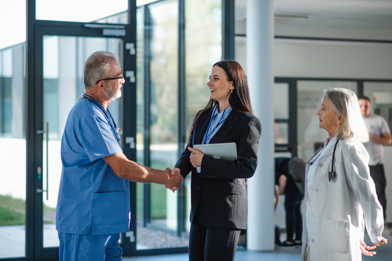 A health care administrator shakes hands with a care provider wearing scrubs in a hospital corridor.
